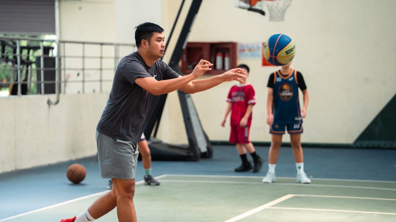 Huấn luyện viên chuyền bóng cho học viên trên sân tập trong nhà, phía sau là các học viên khác