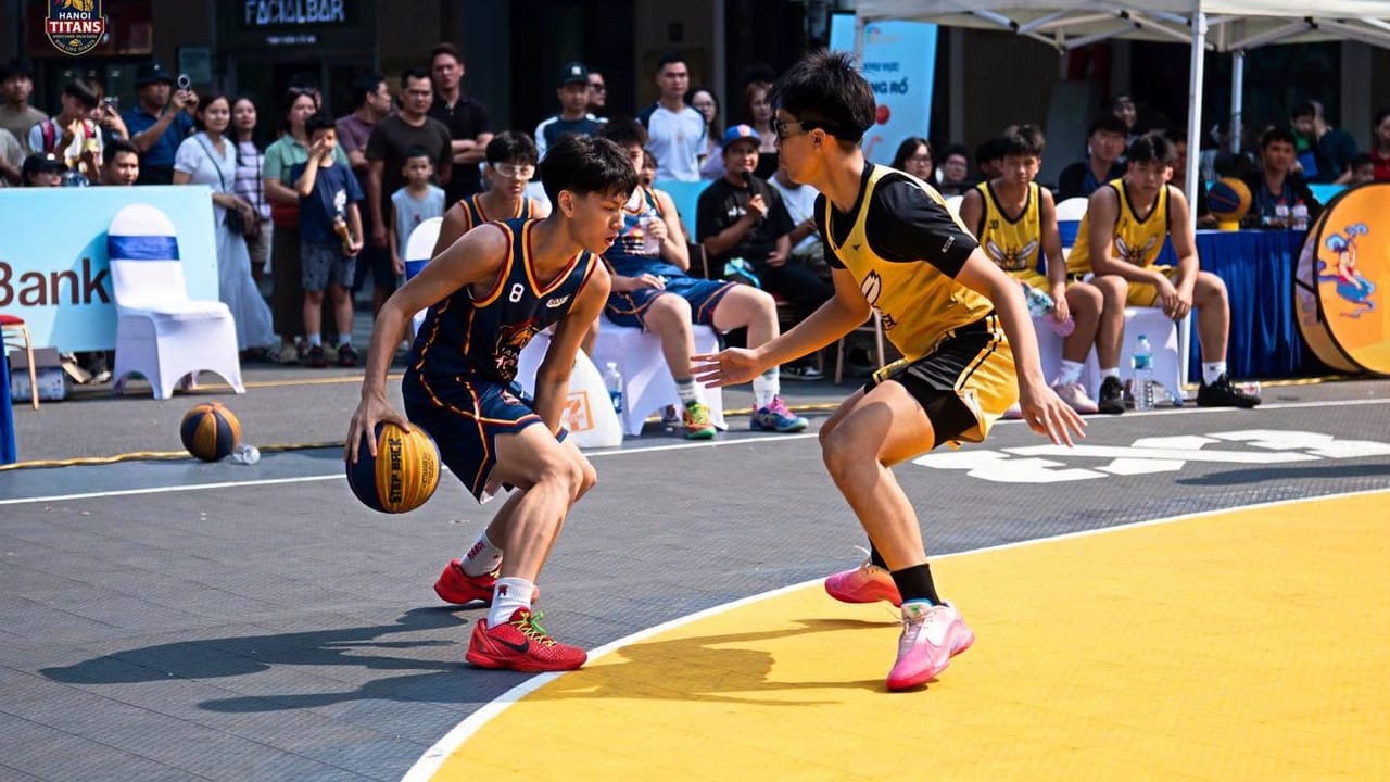 Cầu thủ Hanoi Titans áo xanh navy dẫn bóng trước hậu vệ áo vàng trên sân 3x3 ngoài trời, khán giả đứng xem phía sau