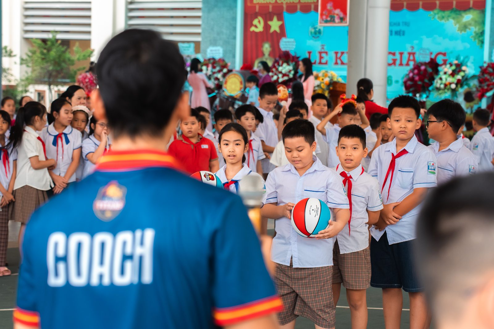 Huấn luyện viên mặc áo in chữ COACH quay lưng, phía trước là học sinh xếp hàng cầm bóng rổ