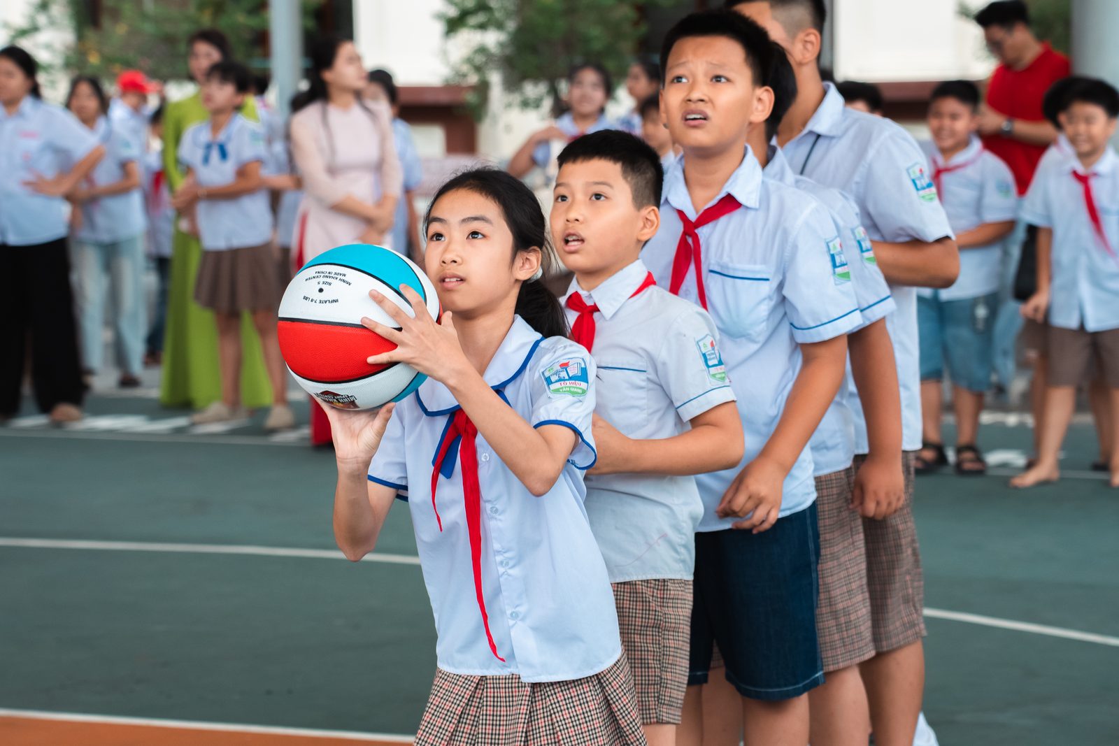 Một nữ sinh tiểu học mặc áo trắng khăn quàng đỏ hai tay nâng quả bóng rổ chuẩn bị ném