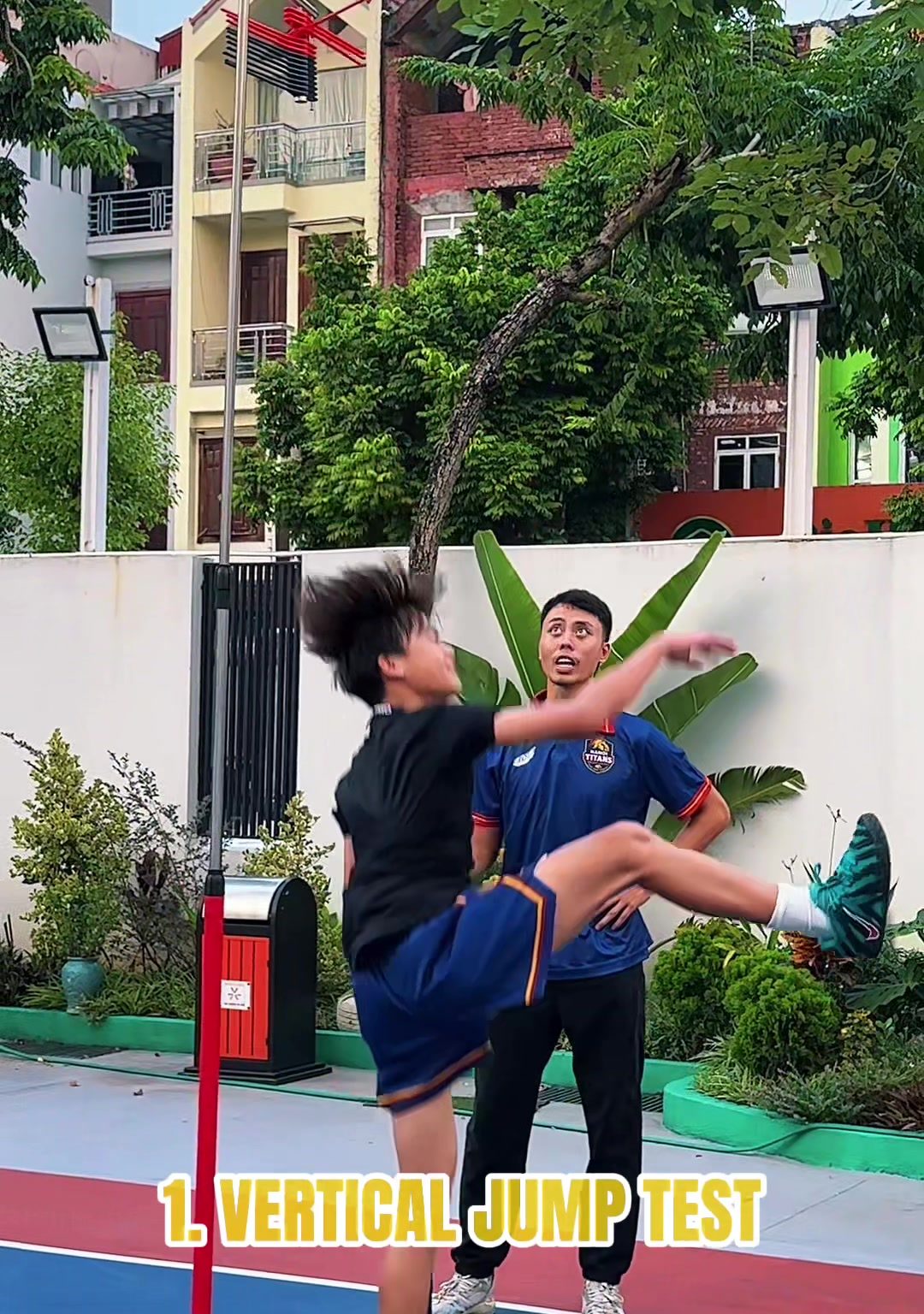 Một học viên bật nhảy cao bên cột đo, huấn luyện viên đứng phía sau quan sát