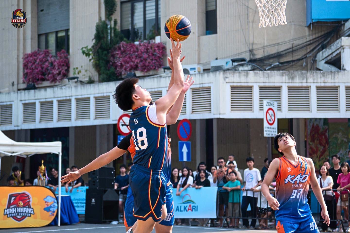 Cầu thủ Hanoi Titans áo số 18 bật lên ném bóng, cầu thủ đối phương áo cam giơ tay chắn, khán giả xếp hàng sau rào chắn