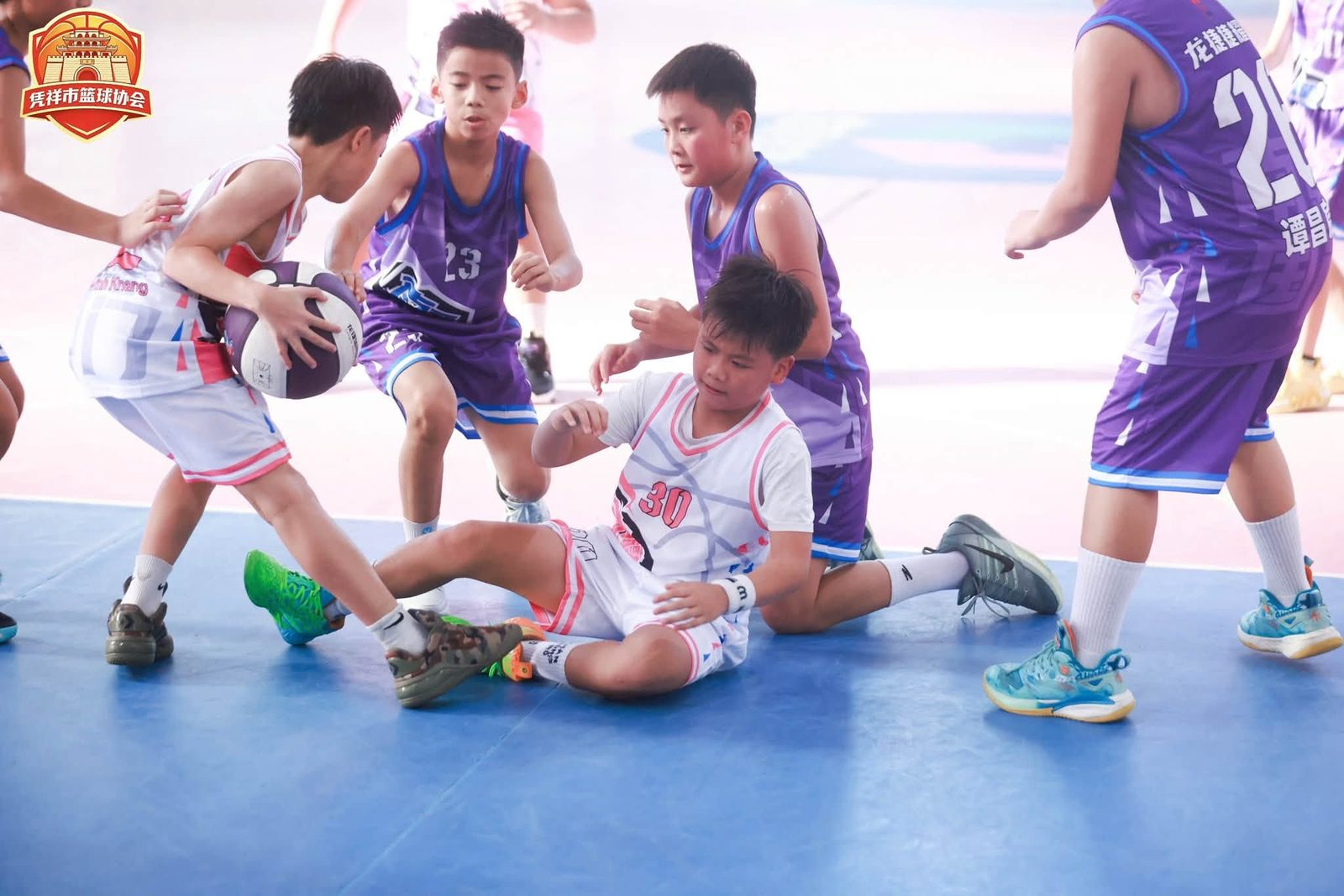 Một cầu thủ nhí áo trắng ngã trên sàn giữa các cầu thủ áo tím đang tranh bóng
